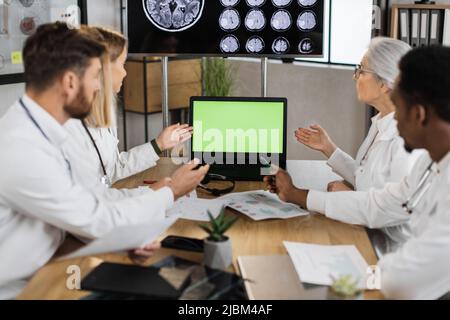 Teamwork of four multiethnic doctors wearing white lab coats and pointing on laptop with green screen while working in polyclinic boardroom. Concept of effective team cooperation in healthcare field. Stock Photo