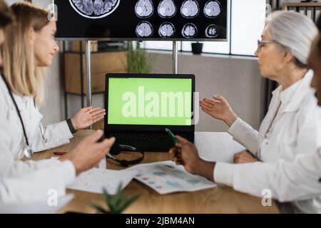 Teamwork of four multiethnic doctors wearing white lab coats and pointing on laptop with green screen while working in polyclinic boardroom. Concept of effective team cooperation in healthcare field. Stock Photo