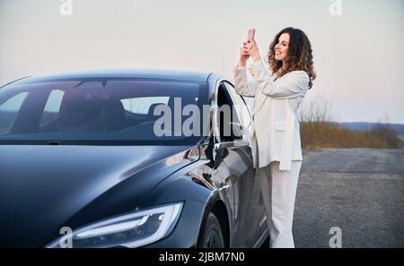 Beautiful curly woman with cute smile standing near her black luxury electric car on road, and taking picture at phone on the backdrop of grey sky. Stock Photo