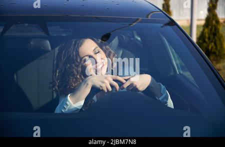 Beautiful businesswoman with closed eyes sitting in electric car and smiling while using vehicle autopilot system. Pretty woman holding steering wheel and sleeping while sitting on driver seat in car. Stock Photo