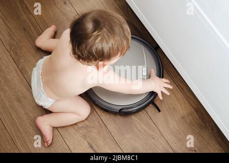 Happy toddler baby boy is playing with a robot vacuum cleaner on the floor. Kid aged one year Stock Photo