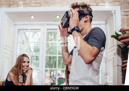 Young man trying virtual reality glasses while people looking at him and laughing in restaurant. Stock Photo