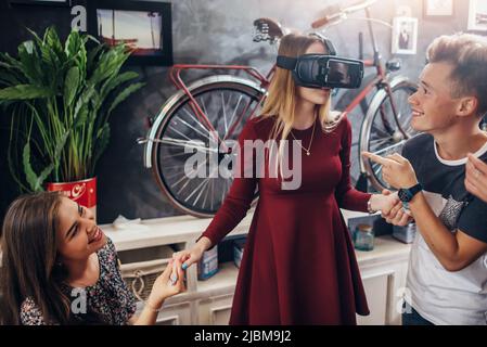 Group of cheerful students supporting their female friend playing funny game using virtual reality helmet in living room. Stock Photo