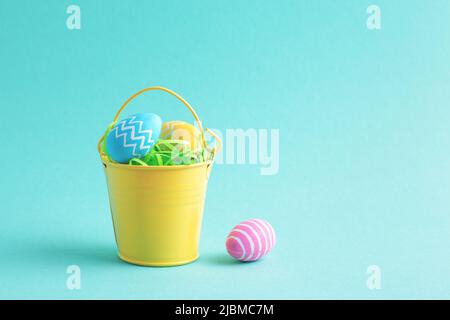Decorative holiday composition with colourful Easter eggs in bright yellow bucket isolated on blue background with copy space. Front view. Stock Photo