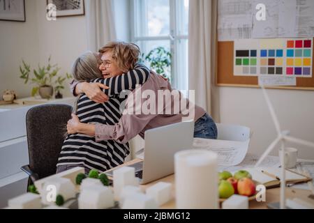 Senior women eco architects with blueprints working on laptop together in office, hugging and having success. Stock Photo