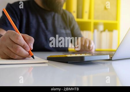 Using laptop and taking notes, close up of man's hand using laptop and taking notes. Selective focus to writing hand, using calculator. Working hard. Stock Photo