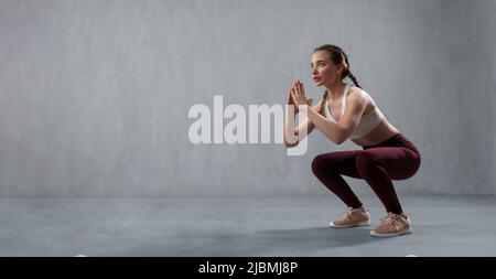 Sports woman in fashion sport clothes squatting doing sit-ups in gym, over gray background Stock Photo