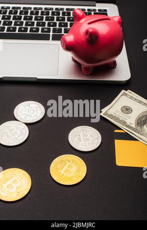 KYIV, UKRAINE - APRIL 26, 2022: High angle view of bitcoins near dollars and blurred laptop on black background Stock Photo