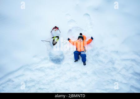 Boy in sport winter orange coat lay holding hand of snowman Stock Photo