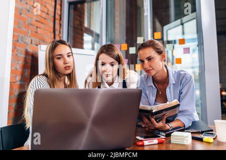 Three female college students studying together using laptop and lecture notes sitting at desk in study room. Stock Photo
