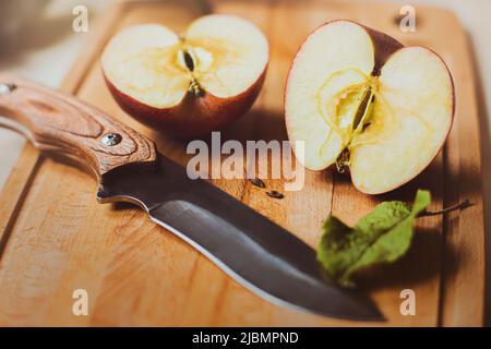 One ripe red sweet apple is lying on a cutting board, cut in half, and a sharp knife is lying next to it. Fruit on the kitchen table. Harvest. Stock Photo