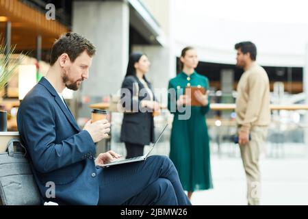 Side view portrait of bearded businessman using laptop and enjoying coffee while taking break in lounge area of office building, copy space Stock Photo
