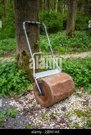 Old rusty garden roller leaning up against a tree in a wooded area. Stock Photo
