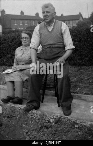 1940s, historical, on a layer of concerete in a back garden, an elderly working class couple sitting outside on stools, the gentleman sitting on a taller stool, hence he looks quite alot larger than the lady, England, UK.. The gentleman is smoking a pipe and wearing a sleeveless sweater over a collarless shirt with rolled up sleeves. Stock Photo