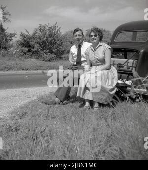 1950s, historical, beside a country road, on a grassy lay-by, a couple sitting on the boot lit of their car, resting, having a break, England, UK. In the open rear boot (or trunk) space can be seen the bulky luggage cases of the era that people used when travelling. Stock Photo