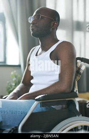 African American man in eyeglasses with disability sitting in wheelchair and looking away with pensive expression Stock Photo
