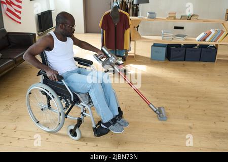 African man with disability sitting in wheelchair and cleaning floor in room using vacuum cleaner without help Stock Photo
