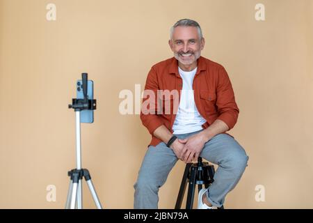 A man in a red jacket sitting on a stool in a studio and making selfie Stock Photo