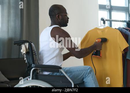 African young man sitting in wheelchair using steamer to iron clothes hanging on the rack Stock Photo