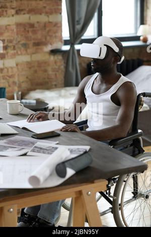 African American man sitting in wheelchair at desk using virtual reality goggles to learn architectural projects Stock Photo