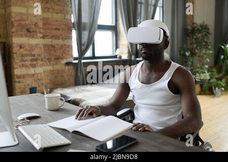 African young man with disability studying online at table using virtual reality headset during rehabilitation at home Stock Photo