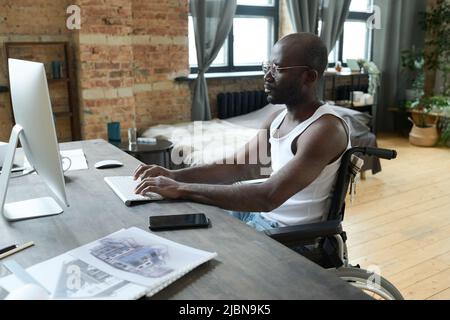 African serious man with disability sitting in wheelchair at table with computer and typing on keyboard, he working over online project Stock Photo