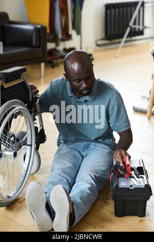 African young man with disability sitting on floor and taking tools from box to repair his wheelchair himself Stock Photo