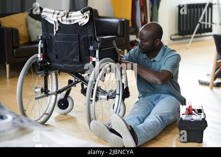 African man with disability sitting on floor and fixing his broken wheelchair with instruments Stock Photo