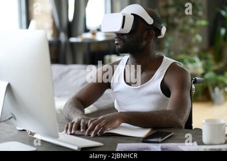 African young man in virtual reality goggles sitting at table in front of computer monitor and typing on keyboard Stock Photo