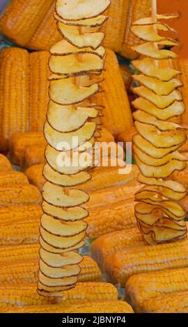 long skewers of fried potato chips with peel and yellow cooked organic corn cobs with plastic wrap for sale in street food stall Stock Photo