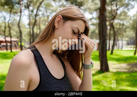 Beautiful brunette woman wearing sports bra standing on city park, outdoors rubbing nose and eyes feeling fatigue and headache. Massaging nose bridge, Stock Photo