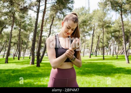 Brunette sportive woman wearing black sports bra standing on city park, outdoors holding her painful wrist. Suffering pain on hands and fingers, arthr Stock Photo