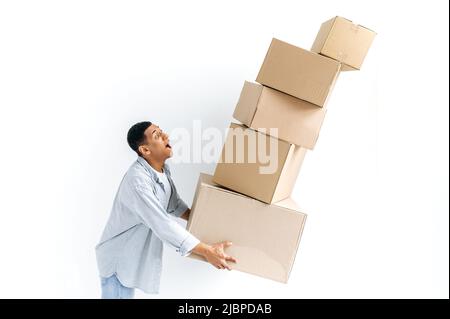 Stunned mixed race guy in casual stylish clothes, delivery man, standing over isolated white background, holding many cardboard boxes, delivering parcels, looking in amazement at falling boxes Stock Photo