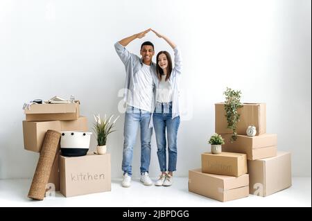 Multiracial excited couple in love, young family, standing near a white wall, between cardboard boxes, in their new apartment, depicts the roof with their hands, looking at camera, smiling happily Stock Photo