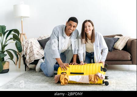 Joyful family multiracial young couple, hispanic guy and caucasian girl, sit on the floor at home in the living room near a large yellow suitcase, putting clothes in a suitcase for vacation, smiling Stock Photo