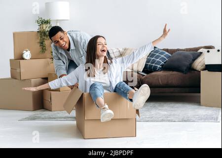 New home, relocation. Happy mixed race young family couple, bought their own house, they having fun on moving day, joyful wife sitting in cardboard box and husband push her, they laughing together Stock Photo
