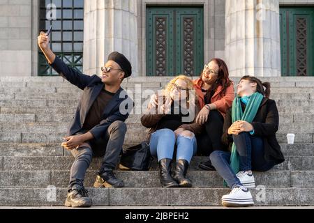 Multi ethnic group of friends sitting on some stairs having a good time chatting, laughing and taking photos Stock Photo
