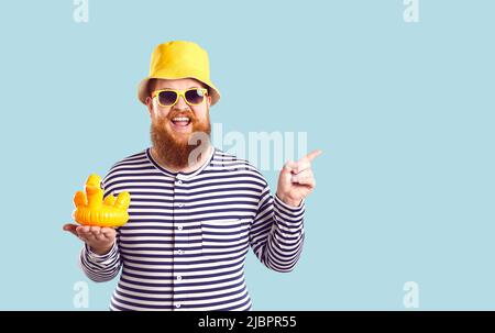 Funny fat man with inflatable duck in his hand points to copy space on pastel light blue background. Stock Photo