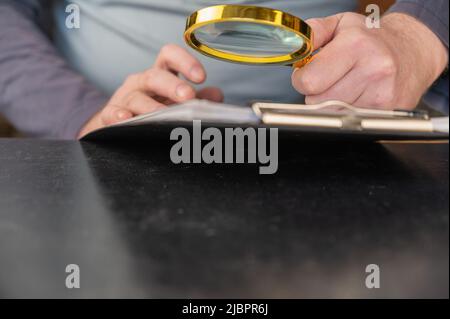 A man's hand holds a magnifying glass over a document. Round magnifying glass with gold-colored frame. Paper clipboard. Inside the room. Selective foc Stock Photo