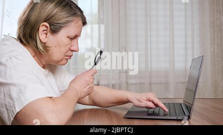 Old woman uses magnifying glass for typing on keyboard Stock Photo