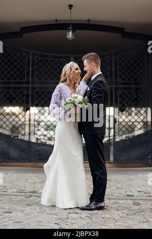 Beautiful wedding couple in elegant suit and dress walk around city, smile and hug on street background. Portrait of happy bride with bouquet and Stock Photo