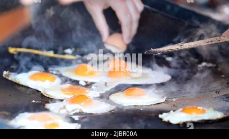 A number of fresh raw eggs sizzling away on a smoking hot black bbq cook top. An egg is cracked in the background to be cooked. Eggs on the barbecue s Stock Photo