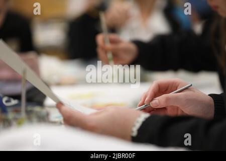 A close up of an art students hand holding a small grey water color paint brush painting at a desk in a school art class room Other students painting Stock Photo