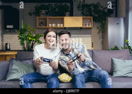Young married couple man and pregnant woman together have fun sitting on sofa and playing electronic games Stock Photo