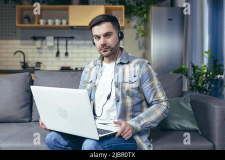 A man works at home remotely, sits on a sofa and uses a laptop and a headset for video calling Stock Photo