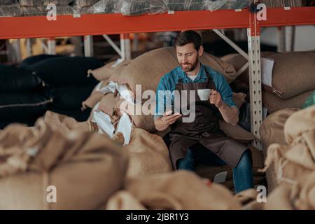 Business owner with cup of coffee and phone sit on warehouse of coffee factory Stock Photo