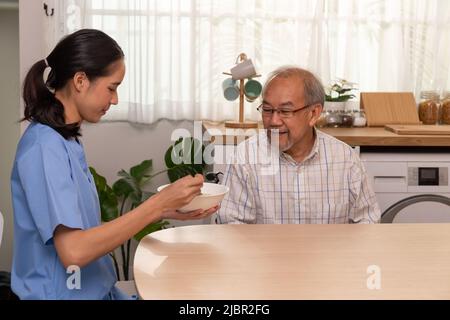 A young Asian nurse take care for a elderly man at retired nursing home service. A caregiver support elderly in a kitchen during breakfast. Stock Photo