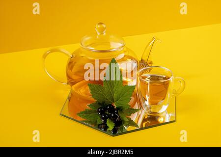 Black currant tea. Transparent glass teapot, cup of tea and black currant berries on a yellow background. Calming drink concept. Stock Photo