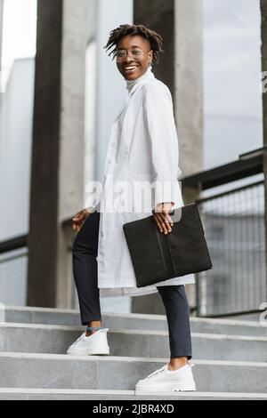 Pleasant young African American woman doctor in white gown, holding a folder, standing on stairs of modern clinic outdoors, smiling and looking at camera. Stock Photo