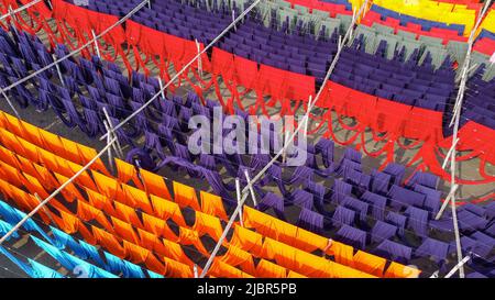 June 8, 2022, Narayanganj, Dhaka, Bangladesh: Workers hang thousands of meters of different colored fabrics to dry so they are able to be made into t-shirts and vests. Iron wires are used between bamboo frameworks to create giant washing lines for the final part of the dying process as the fabrics are dried in the sun in Narayanganj, Bangladesh. Bright strands of blue, pink, orange, red, purple, yellow, magenta and green-dyed cloths hang above the grassy field in a dazzling network of interlocking colors. After five to six hours in the sun, the fabric is used to make dresses.This is the final Stock Photo